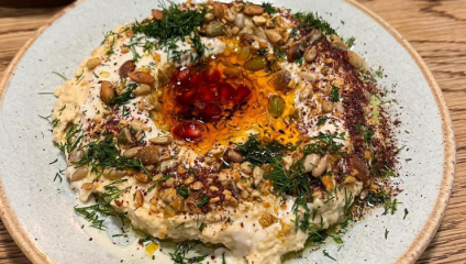 A plate of creamy hummus is garnished with herbs, seeds, a sprinkle of spices, and a drizzle of oil. A pool of red sauce is in the center of the hummus, adding a vibrant touch. The dish is presented on a rustic wooden table.