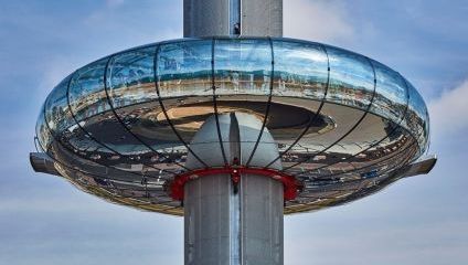 A close-up view of a futuristic glass observation tower pod, circular in shape, reflecting the surrounding scenery. The pod is attached to a tall central column and appears elevated above the ground against a blue sky backdrop.