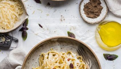 Two bowls of creamy pasta are displayed on a white surface with forks resting in each bowl. Around them are small dishes containing grated cheese, ground black pepper, and olive oil. Fresh herbs are scattered for garnish, along with two beige cloth napkins.