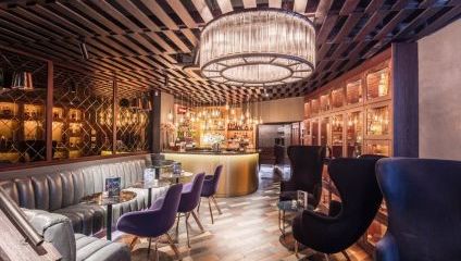 A modern, stylish bar with a circular counter illuminated by a grand chandelier above. The setting includes dark velvet chairs, cushioned sofas, and small round tables. The decor features wooden slats on the ceiling and elegant lighting, creating a cozy atmosphere.