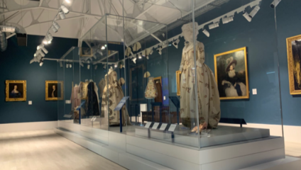 A museum exhibit displays mannequins wearing historical dresses behind glass cases. Framed paintings of various individuals hang on the deep teal walls. Overhead spotlights illuminate the scene, highlighting the intricate details of the clothing and artwork.