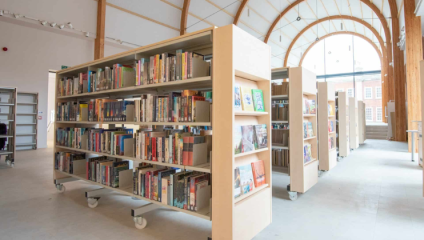 A spacious library interior with tall, wooden arches and large windows. Bookshelves filled with various books and magazines are arranged in rows, each on wheels for mobility. The space is bright and neatly organized, creating a serene atmosphere for reading.