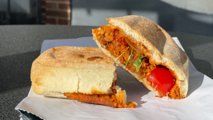A close-up of a sandwich made with ciabatta bread, filled with visible ingredients like red bell peppers, leafy greens, and a reddish-orange spread. The sandwich is cut in half, with one half placed on its side, revealing the filling.