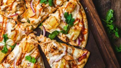 A close-up of a pizza topped with cheese, pineapple, red onions, shredded chicken, and fresh cilantro. The pizza is sliced into six pieces and sits on a wooden cutting board. Fresh cilantro leaves and a red onion are visible around the cutting board.