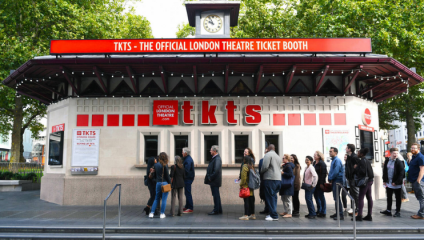 People stand in line at the official TKTS London Theatre Ticket Booth, a white building with a red sign and clock on the roof, located outdoors with trees in the background. The booth features large TKTS letters and additional signage promoting theatre tickets.