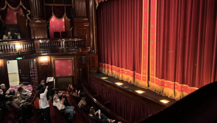 A theater stage with a closed red curtain featuring gold accents. Audience members sit in seats, some on their phones. The balcony has ornate wooden details and pink decorations. Illuminated stage lights highlight the curtain's base.