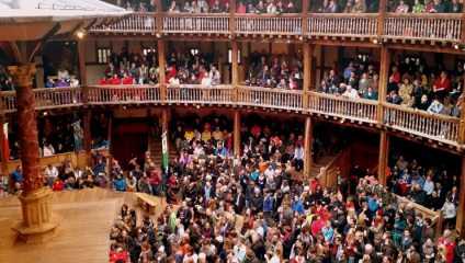 A large audience fills the multi-tiered wooden seating and ground area of a circular theater, reminiscent of Shakespeare's Globe. The theater is packed with people eagerly waiting for a performance with many standing on the ground level in front of the stage.