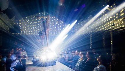 A performer dances on a disco ball under bright, colorful lights in a dim club with a captivated audience seated around the stage. The ceiling and walls are adorned with light reflections, creating an energetic and vibrant atmosphere.