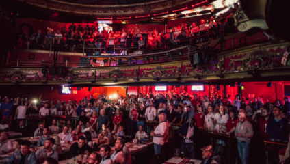 A packed theater with two levels of seated and standing audiences, illuminated by red and warm lights. The ornate interior features intricate decorations, balconies, and railings. Many attendees seem engaged, some taking pictures or recording with their phones.