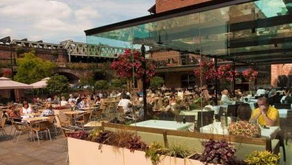 Outdoor restaurant bustling with people dining under partially covered seating areas. Hanging flower baskets and potted plants adorn the glass roof structure. A historical bridge and greenery are visible in the background under a clear sky.