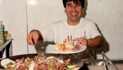 A smiling person stands behind a kitchen counter, holding a plate with food and offering it towards the camera. A pan filled with sliced meat and vegetables is in the foreground. The person wears a white t-shirt and the background features a tiled wall.