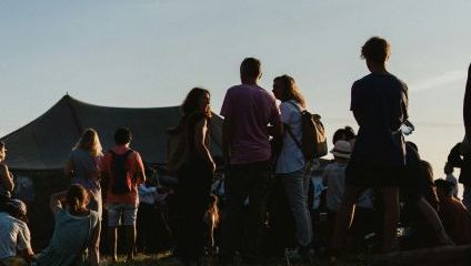 A group of people is gathered outdoors in a grassy area, enjoying an event or festival as the sun sets. Some are standing and chatting, while others are seated on the grass. A large tent is visible in the background under a clear sky.
