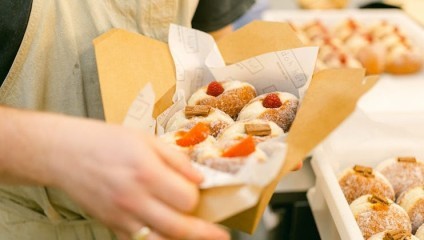 A person holds a brown paper tray filled with sugared doughnuts. The doughnuts are topped with fresh raspberries and strawberries. More doughnuts are visible in trays in the background. The person is wearing a light-colored apron.