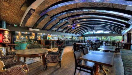 A spacious, stylishly designed restaurant interior with wooden flooring, an arched ceiling featuring blue accents, and a variety of seating including upholstered and wooden chairs and tables. The walls have a mix of exposed brick and contemporary elements.