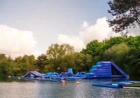 A large inflatable water park sits on a calm lake surrounded by dense, green trees. The park features multiple blue inflatable slides, climbing walls, and obstacle courses. The sky above is partly cloudy, and a few buoys float near the structures.