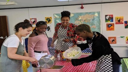 Four people are cooking together in a brightly decorated room. Three are standing around a red polka-dot table, pouring ingredients from a pot, while one person is on the left, laughing. Walls feature colorful art and a world map. Everyone is wearing aprons.