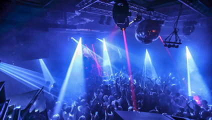 A crowded nightclub with vibrant blue and purple lighting illuminates people dancing. Bright beams of light and lasers cut through the smoky air, while a disco ball hangs from the ceiling, reflecting the colorful lights onto the crowd below.