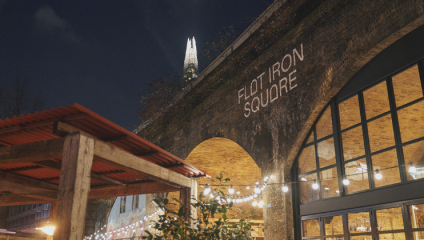 A nighttime view of Flat Iron Square in London, with the Shard skyscraper illuminated in the background. The Square's name is projected onto a brick archway, and string lights adorn the area, adding a warm and festive ambiance.