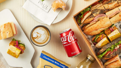 A variety of sandwiches displayed on a light-colored surface. Included are a boxed assortment of sandwiches, a Coca-Cola can, a latte in a clear cup, a white plate with a sandwich, and a paper bag with a pastry. Various condiment packs and a small bottle also present.