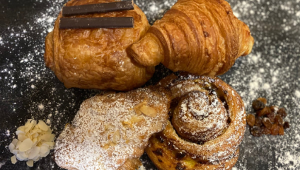 A close-up of assorted pastries on a dark surface dusted with powdered sugar. The assortment includes a pain au chocolat topped with chocolate strips, a croissant, an almond-topped pastry, and a cinnamon roll. Almond flakes and raisins are scattered beside the pastries.