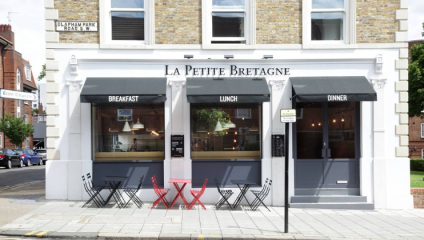 A quaint restaurant named La Petite Bretagne on a street corner, with a beige brick facade and black awnings. Red and black chairs with small tables are set outside. Signage indicates meals are served for breakfast, lunch, and dinner. A street sign reads Clapham Park Road SW4.