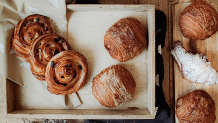 A wooden tray holds several types of pastries, including cinnamon rolls with raisins, croissants, and pain au chocolat. A dusted croissant lies on a smaller wooden tray to the right, adding to the appetizing display of baked goods.