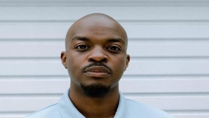 A person with a neutral expression is standing in front of a white, horizontal panel background. They are wearing a light blue shirt and have a shaved head with a short beard and mustache.