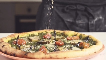 A close-up shot of a person sprinkling cheese onto a freshly baked pizza topped with cherry tomatoes, zucchini slices, and pesto. The pizza is on a pink plate, and the person is wearing a dark apron in the background.