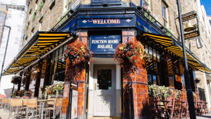 A charming pub with a brick exterior and striped yellow and black awnings. Over the entrance, a sign reads Welcome and Function Rooms Available. Hanging flower baskets and pots add vibrant colors. Outdoor seating with wooden tables and chairs lines the sidewalk.