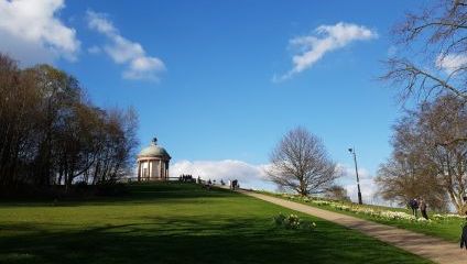 A small, round pavilion with a domed roof sits atop a grassy hill under a clear blue sky with a few scattered clouds. A paved path lined with bare trees and some people walking leads up to the pavilion. Forested areas flank the scene.