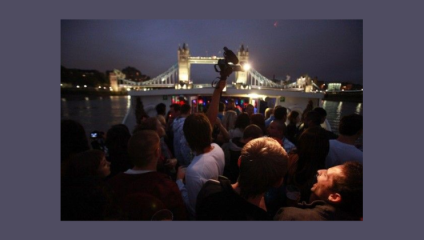 A group of people are gathered on a boat at night, facing towards the brightly lit Tower Bridge in the background. The scene appears lively, with someone holding up a camera to capture the moment. The sky is dark, adding to the vibrant atmosphere.