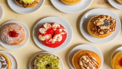 A selection of various doughnuts on white plates arranged on a light wooden surface. The doughnuts are decorated with different toppings such as icing, sprinkles, chocolate drizzle, nuts, and fruit preserves, showcasing an assortment of flavors and styles.