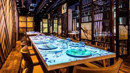A modern restaurant interior with long tables set for dining. The tables have glowing designs, and clear glasses and plates are neatly arranged. Wood chairs surround the tables, and the ambient lighting creates a warm, inviting atmosphere.