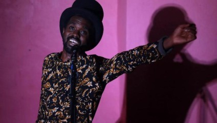 A person wearing a patterned shirt and a dark hat performs enthusiastically in front of a microphone, with a pink wall and their shadow in the background. The individual appears to be singing or speaking, with one arm extended and a joyful expression on their face.