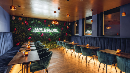 A modern restaurant interior with a neon sign reading Jam Delish Modern Caribbean Vegan Lounge against a plant-covered wall. Teal velvet chairs and wooden tables are arranged neatly, while warm, dim lighting creates a cozy atmosphere.