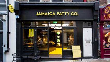 Street view of Jamaica Patty Co., a black-framed storefront with illuminated yellow signage. The entrance showcases a modern interior with bright lighting. A sidewalk table and chair are visible to the left, and a person is inside near the counter.