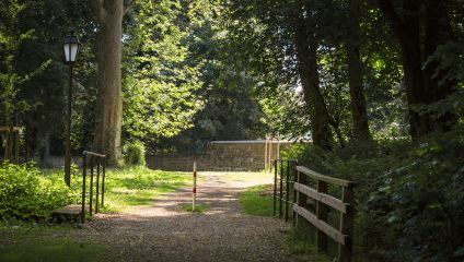 A serene park path surrounded by lush green trees and foliage. The gravel path is lined with a wooden fence on the right and black metal fences on the left, leading towards a stone wall in the distance. A lamp post stands to the left of the path.