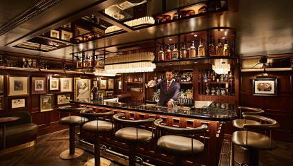 A bartender in a suit is pouring a drink at a luxurious, dimly lit bar with a dark wood interior. The bar has high stools, illuminated shelves with various bottles, framed pictures on the walls, and elegant pendant lights hanging above.