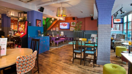 A brightly colored, eclectic restaurant with various seating options, including wooden chairs with patterned cushions and a purple booth. The walls are adorned with art and framed pictures. Natural light streams in through large windows, and a staircase is in the background.