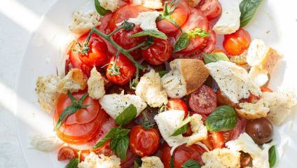 A fresh tomato and mozzarella salad is served on a white plate. The salad includes cherry tomatoes, basil leaves, crumbled bread pieces, and mozzarella chunks, all drizzled with olive oil and seasoned with salt and pepper. Sunlight and shadows add contrast.