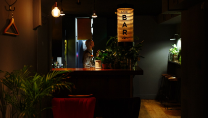 A dimly lit bar with a cozy ambiance is shown. A bartender is standing behind the counter, surrounded by potted plants. A vertical lamp with the word BAR illuminates the scene. There are wooden bar stools lined up at the far end of the counter.