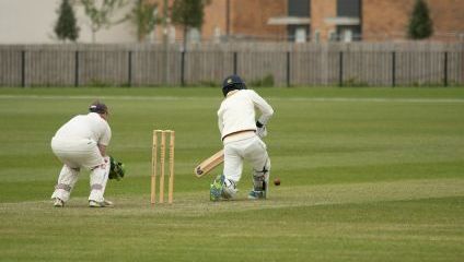 Two cricket players in action. The batsman has just played a shot while the wicketkeeper waits behind the stumps. Both players are dressed in white cricket uniforms. The game is being played on a grassy field, with residential buildings visible in the background.