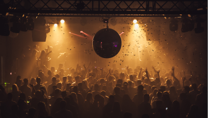 A crowded dance floor filled with people dancing under dim, colorful lighting. A large disco ball hangs from the ceiling, reflecting light. Confetti fills the air, and the atmosphere is lively and energetic with music and celebration.