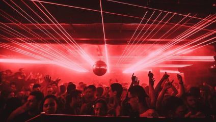 A crowded nightclub filled with people dancing under red lighting and laser beams. A silver disco ball hangs from the ceiling, reflecting the vibrant lights. The atmosphere is energetic and lively, with hands raised in the air and visible excitement among the crowd.