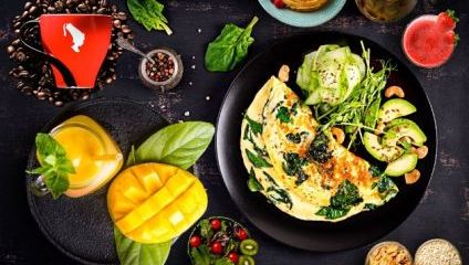 A vibrant breakfast spread on a dark table. It includes an omelette with greens, sliced avocado, cucumber salad, a sliced mango, pancakes, a glass of orange juice, a strawberry drink, a red coffee cup, spices, and a small bowl of nuts and seeds.