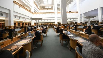 A spacious library reading room filled with long wooden desks and individual study lamps. People, some using laptops and others with books, are seated at the desks. Shelves of books surround the room, and the high ceiling and large windows allow natural light to flood in.