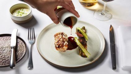 A hand is pouring sauce onto a gourmet dish consisting of a rolled meat entrée topped with crumbled cheese, accompanied by grilled asparagus. The plate is set on a white tablecloth with a fork, knife, bowl of sauce, bread plate, and glasses of wine nearby.