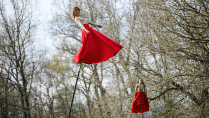 Two performers dressed in flowing red dresses balance on tall poles in a forest. The trees around them are bare, suggesting it is early spring or late autumn. The performers appear to be gracefully moving, creating a captivating scene against the natural backdrop.