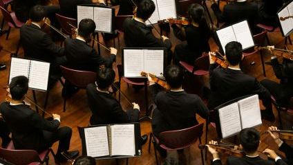 An overhead view shows a large group of musicians in black formal attire seated in rows, playing string instruments and reading sheet music on stands. They are performing on a wooden stage with maroon chairs. The musicians' faces are mostly turned away from the camera.