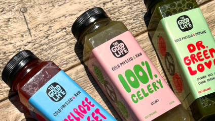 Three bottles of cold-pressed juice from The Good Life are displayed on a rustic wooden surface. The flavors visible are Primrose Sweet, 100% Celery, and Dr. Green Love. Each bottle has a different, colorful label, indicating their unique ingredients.